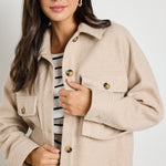 A woman wearing a taupe oversized shacket with a button front, structured collar, and patch pockets, styled with a striped knit top and dark denim jeans.