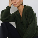 A woman wearing a dark khaki knit cardigan with oversized buttons, styled with black jeans and gold statement earrings.