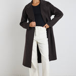 Woman wearing a chocolate brown longline coat over a black tank and cream wide-leg jeans, styled with black boots and sunglasses.