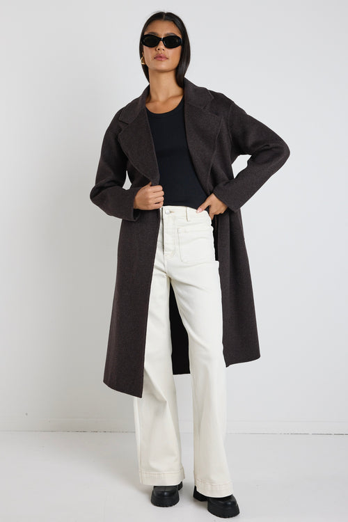 Woman wearing a chocolate brown longline coat over a black tank and cream wide-leg jeans, styled with black boots and sunglasses.