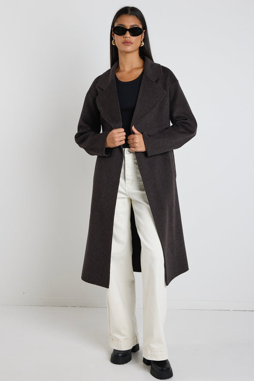 Woman wearing a chocolate brown longline coat over a black tank and cream wide-leg jeans, styled with black boots and sunglasses.
