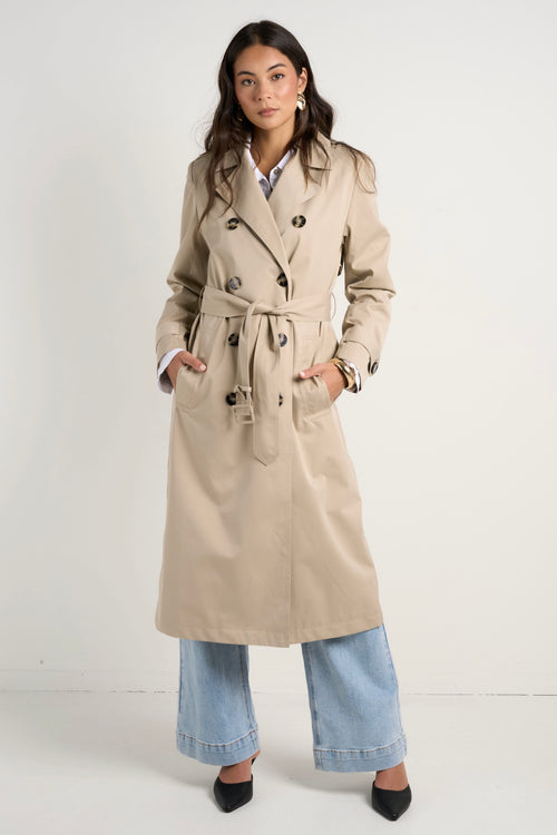 model wears a long beige trench coat with buttons and a collar paired with blue jeans and a white shirt 