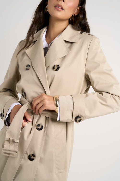 model wears a long beige trench coat with buttons and a collar paired with blue jeans and a white shirt 