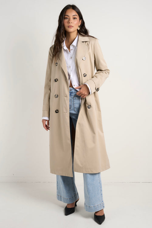 model wears a long beige trench coat with buttons and a collar paired with blue jeans and a white shirt 