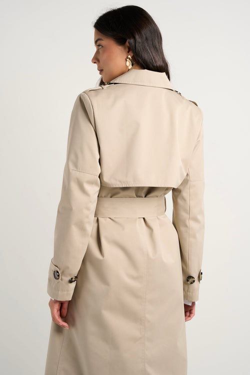 model wears a long beige trench coat with buttons and a collar paired with blue jeans and a white shirt 