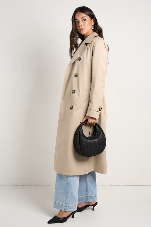 model wears a long beige trench coat with buttons and a collar paired with blue jeans and a white shirt 