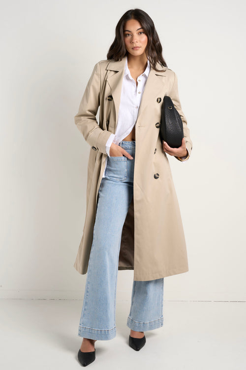 model wears a long beige trench coat with buttons and a collar paired with blue jeans and a white shirt 