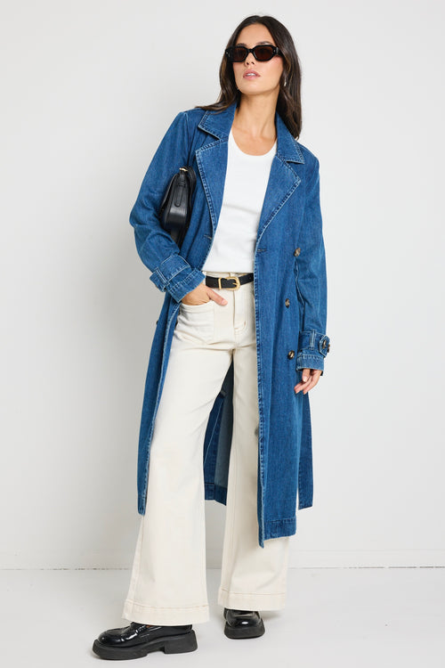 Model wears blue denim trench coat with tie waist and buttons