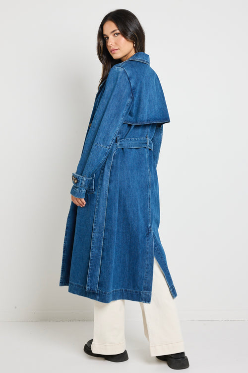 Model wears blue denim trench coat with tie waist and buttons