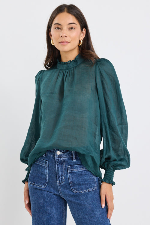 Woman wearing a forest green oversized long-sleeve top with shirred cuffs and a subtle frill neckline, paired with blue jeans.