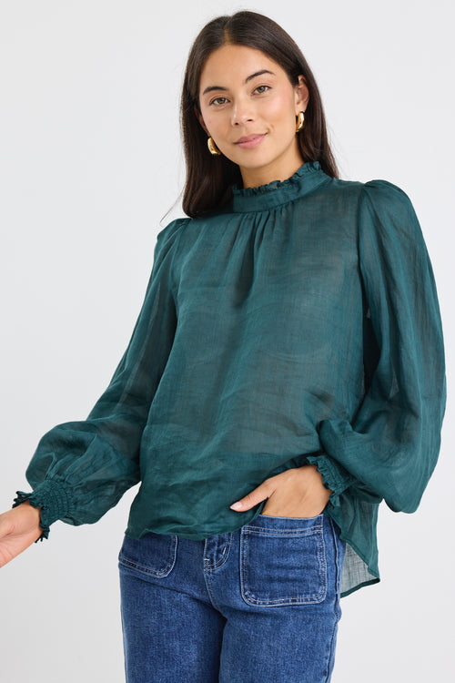 Woman wearing a forest green oversized long-sleeve top with shirred cuffs and a subtle frill neckline, paired with blue jeans.