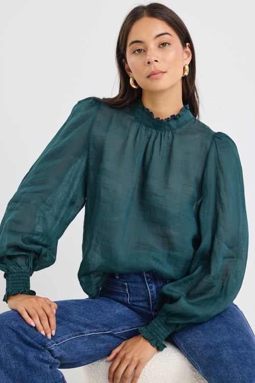 Woman wearing a forest green oversized long-sleeve top with shirred cuffs and a subtle frill neckline, paired with blue jeans.