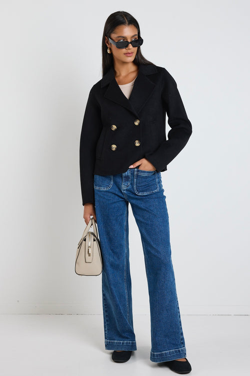 A woman wearing a black marle wool-blend cropped coat with gold buttons, styled with a beige tank top and blue jeans.