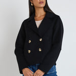 A woman wearing a black marle wool-blend cropped coat with gold buttons, styled with a beige tank top and blue jeans.