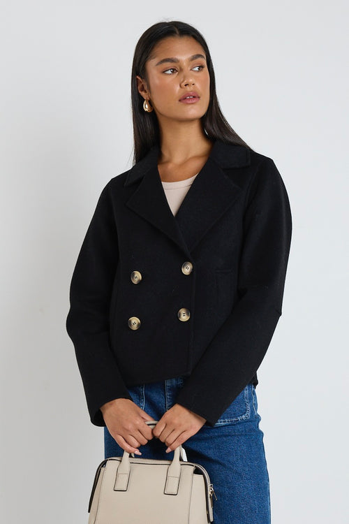 A woman wearing a black marle wool-blend cropped coat with gold buttons, styled with a beige tank top and blue jeans.
