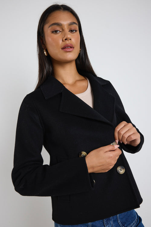 A woman wearing a black marle wool-blend cropped coat with gold buttons, styled with a beige tank top and blue jeans.