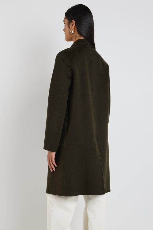 Woman wearing a long khaki coat with tort buttons and wide-leg ecru jeans, looking over her shoulder in profile.







