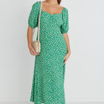 Model wears a green floral print maxi dress with white sneakers and a beige handbag.  