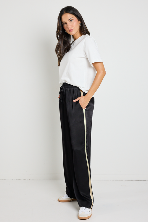 Woman wearing black Townie satin pants with a sandy beige side stripe, paired with a white tee for a sporty yet sophisticated look.