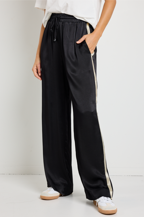 Woman wearing black Townie satin pants with a sandy beige side stripe, paired with a white tee for a sporty yet sophisticated look.