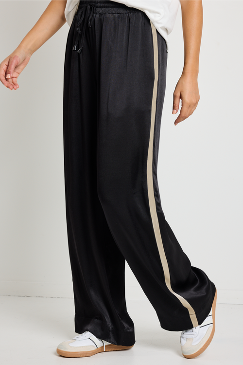 Woman wearing black Townie satin pants with a sandy beige side stripe, paired with a white tee for a sporty yet sophisticated look.