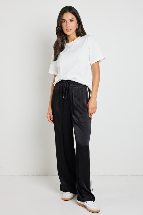 Woman wearing black Townie satin pants with a sandy beige side stripe, paired with a white tee for a sporty yet sophisticated look.