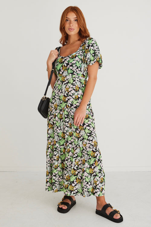 model wearing green and black floral dress and black sandals holding black handbag