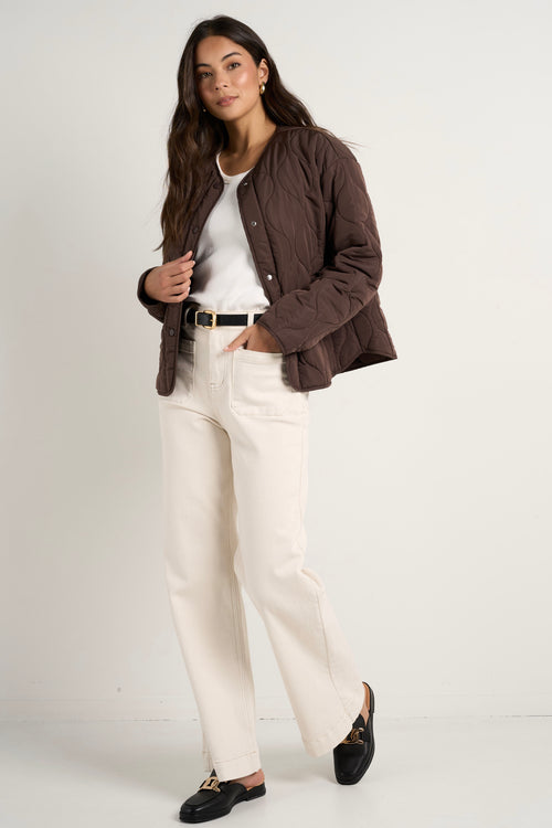 model wears a chocolate brown collarless puffer jacket with  cream jeans