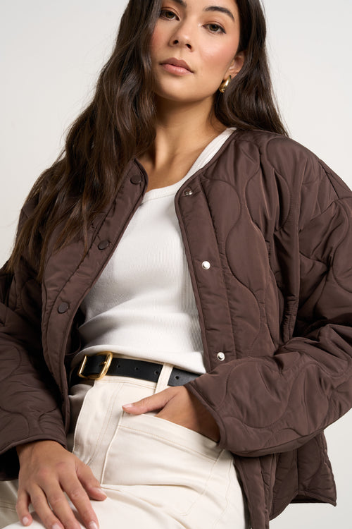 model wears a chocolate brown collarless puffer jacket with  cream jeans